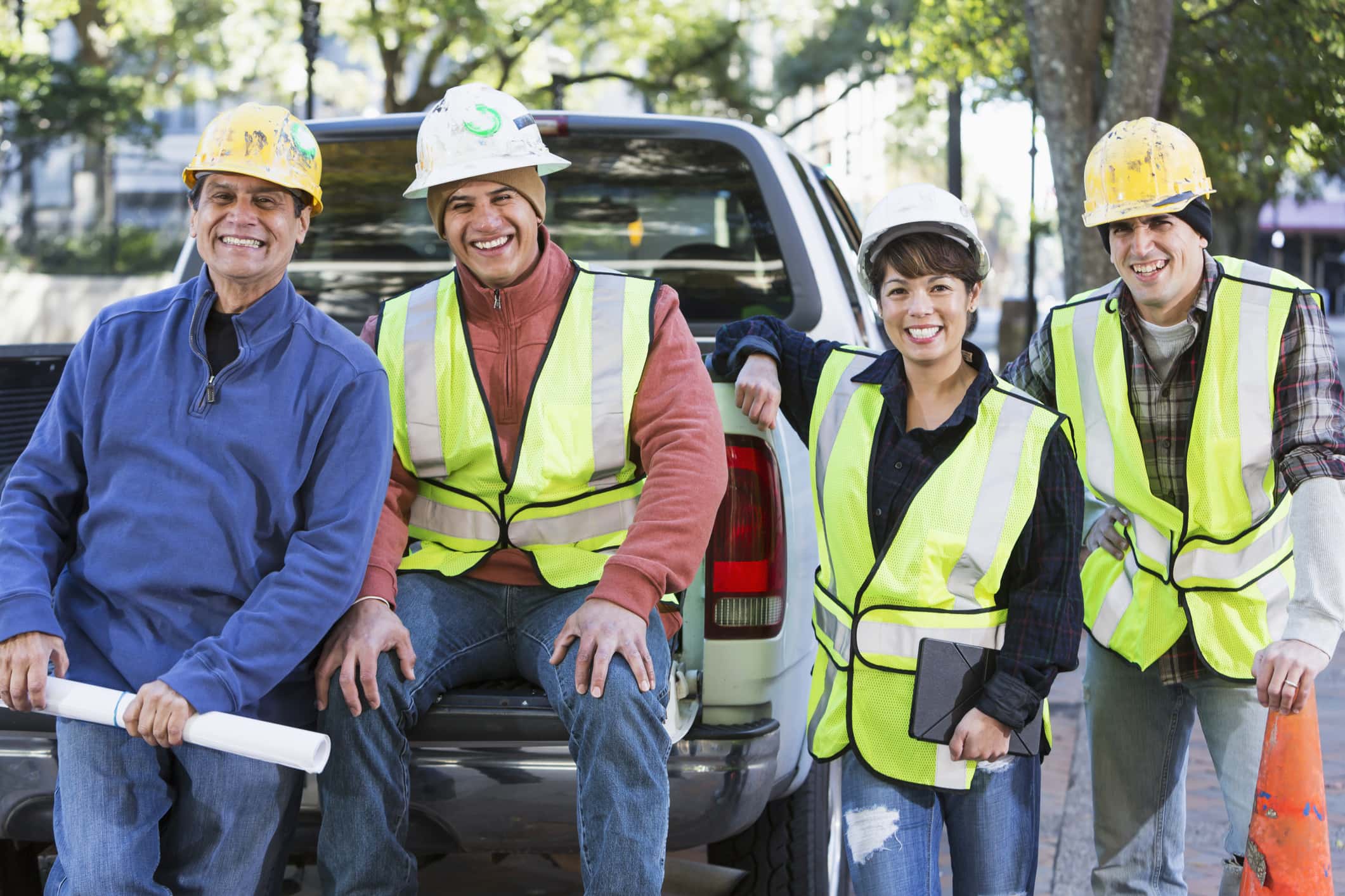 A group of four diverse workers wearing hard hats and yellow safety vests with pickup truck on city street.