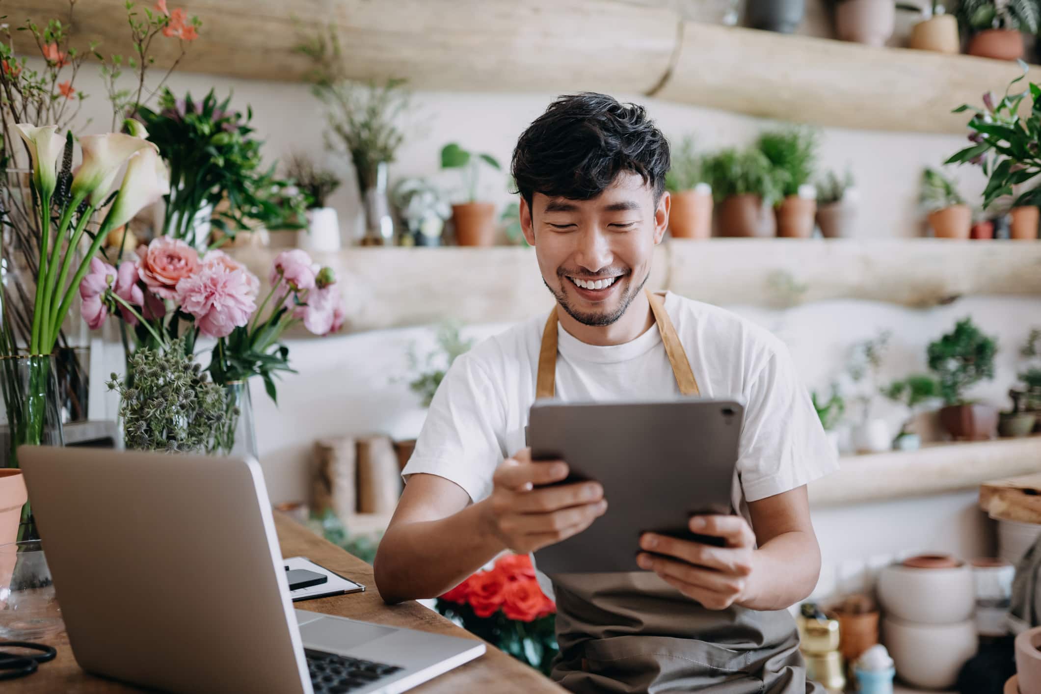 Asian male florist, owner of small business flower shop, using digital tablet while working on laptop against flowers and plants.