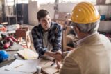 Young smiling carpenter shaking hand with quality control inspector in a workshop.