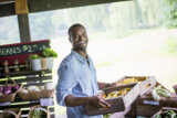 An organic fruit and vegetable farm stand at a farmer's market. A man carrying vegetable boxes.